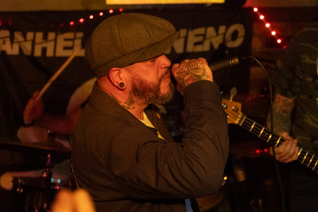 Vocalist in a flat cap singing in front of the Anhell Veneno banner